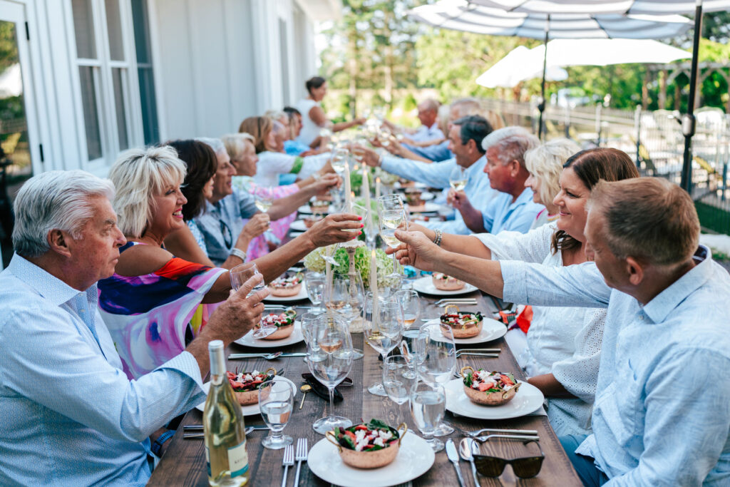 Group gather around a farmhouse table for dinner holding wine glasses up for a toast
