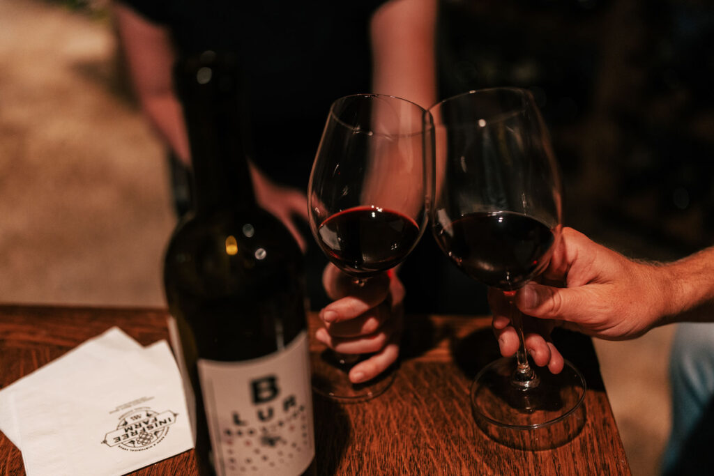 Two hands each holding glasses of red wine in the Inisfree Estate wine cellar.